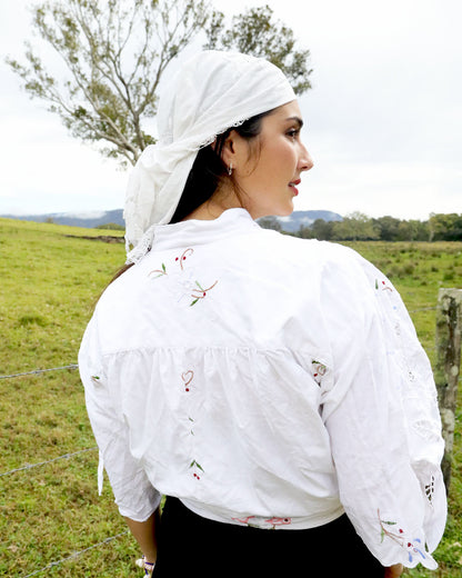 A woman wearing a white embroidered blouse and matching white headscarf, photographed outdoors in a grassy field with a tree and fence in the background.