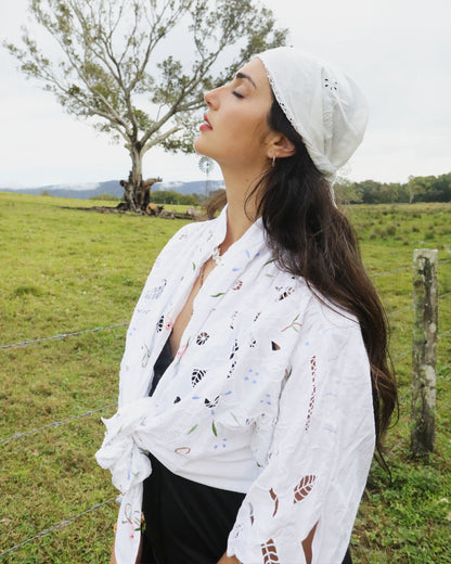 A woman wearing a white embroidered signature Dear Abigael heirloom one size plus size shirt and matching headscarf stands outdoors in a grassy field with a tree in the background.