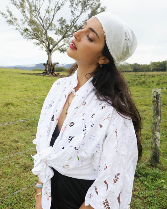 A woman outdoors in a grassy field wearing a plus size, one size, signature Dear Abigael heirloom white embroidered eyelet shirt tied at the waist, a white headscarf, and a black top.