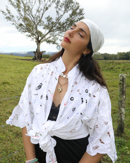 A woman outdoors in a grassy field wearing a white eyelet shirt tied at the waist, a white headscarf, and a black top. A tree and fence are visible in the background.