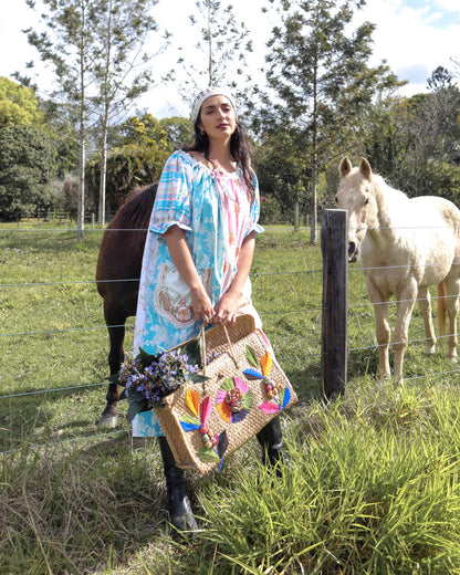 A woman stands in a grassy field with two horses behind a wire fence, holding a large woven straw tote bag decorated with colorful floral embroidery and tassels.