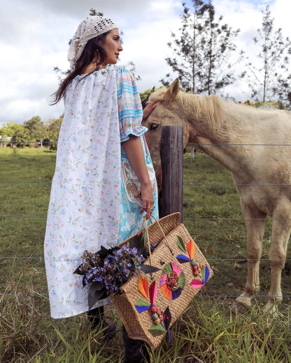 A woman in a white floral cape and crochet head covering stands beside a horse in a grassy field, holding a woven straw tote bag decorated with colorful embroidered flowers and leaves.