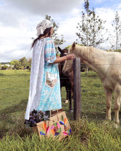 A woman in a long light blue floral dress and white headscarf stands beside a wooden fence in a grassy field, touching a white horse. A woven tote bag with colorful floral embroidery rests on the ground near her.