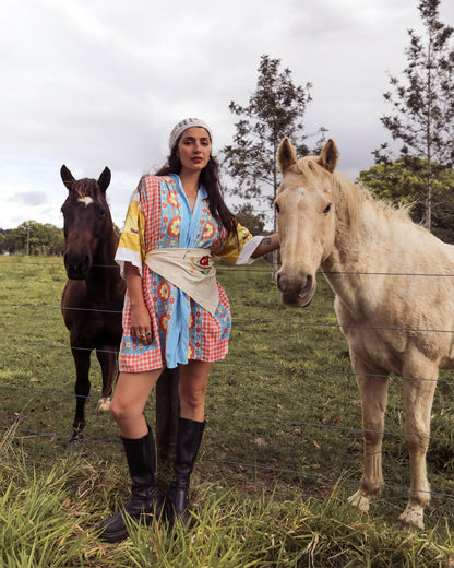 A woman wearing a colorful bohemian-style dress and black boots stands in a grassy field between two horses, one dark brown and one light cream.