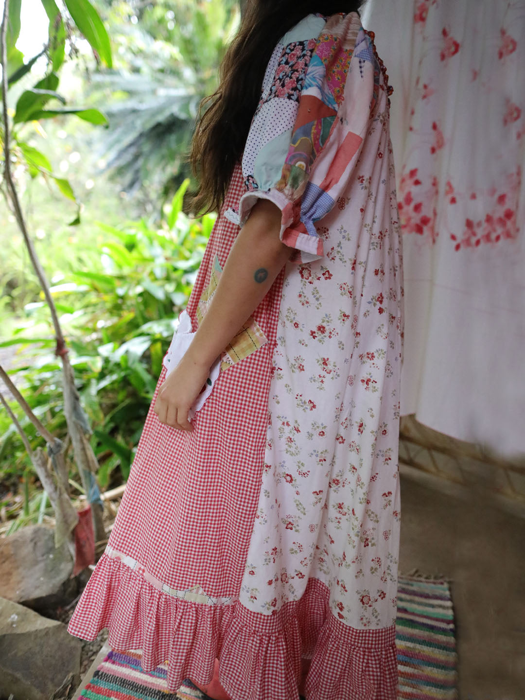 A woman wearing a bohemian patchwork maxi dress with red gingham, white floral prints, and colorful patterned details, standing in a garden setting.