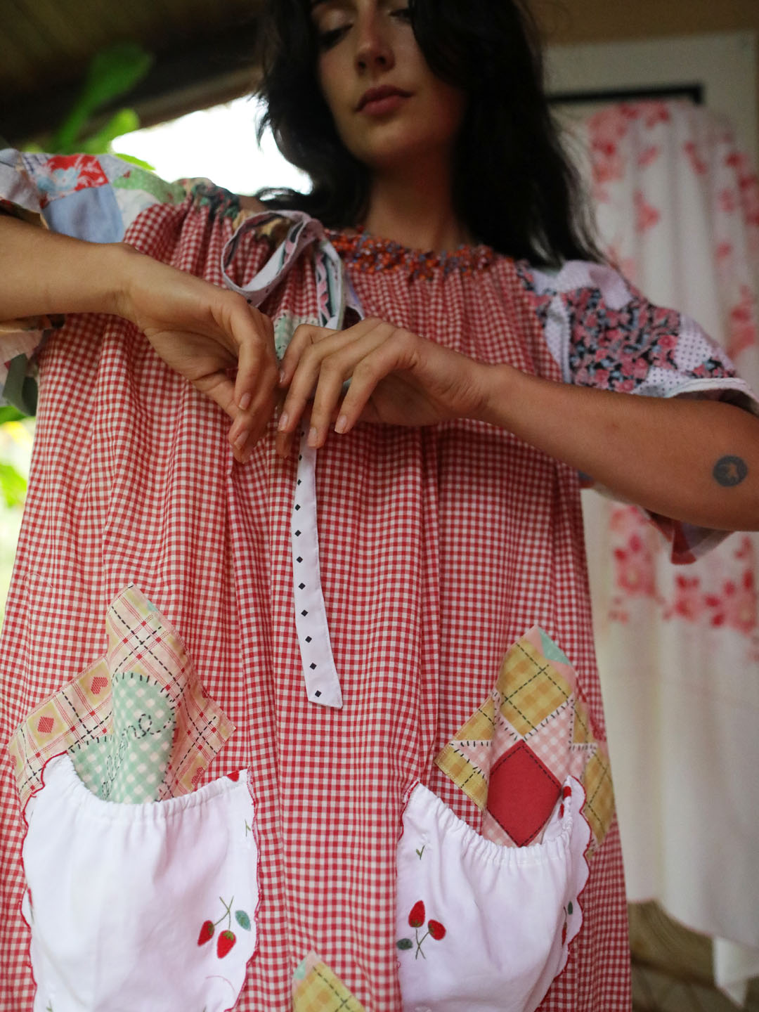 A woman wearing a one size bohemian style red and white gingham patchwork smock dress with white pockets featuring strawberry embroidery and quilted patchwork sleeves.