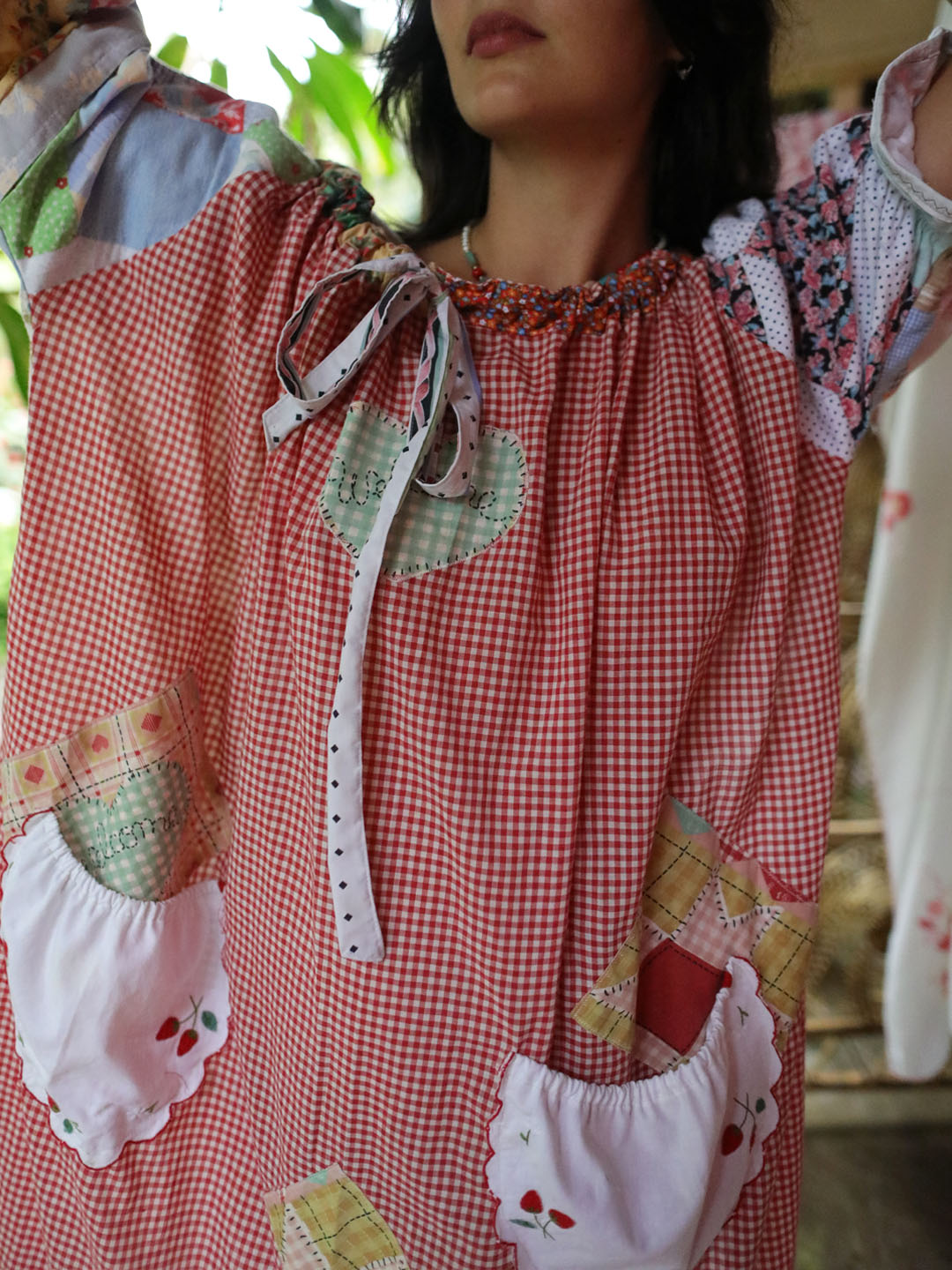 A close-up of a person wearing a patchwork-style red and white gingham dress with floral sleeves, heart-shaped patchwork details, and white ruffled pockets with embroidered strawberries.