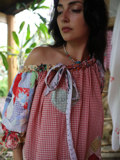 A woman wearing a red and white gingham patchwork top with off-the-shoulder straps, a heart-shaped patch detail, and a tie-neck ribbon, photographed in a rustic outdoor setting.