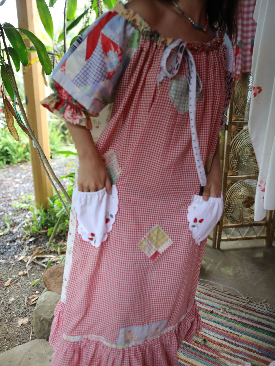 A person wearing a pink and white gingham patchwork dress with ruffled sleeves, tie neckline, and white ruffled pocket flaps with small red embroidered details, photographed in an outdoor rustic setting.