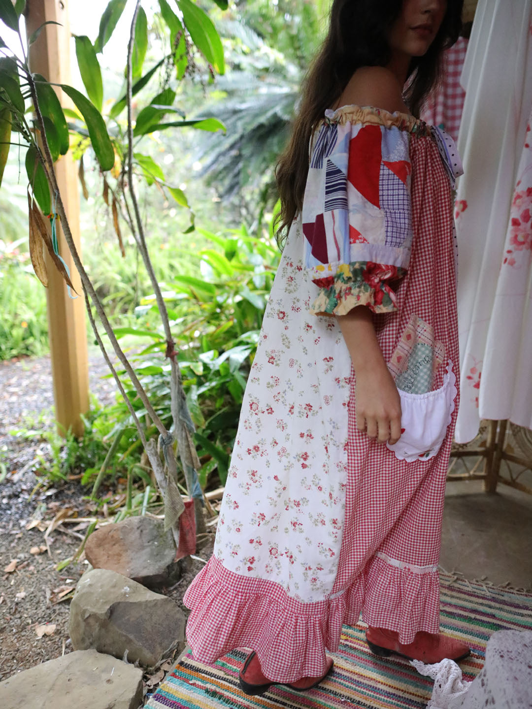 A woman wearing a handmade patchwork off-the-shoulder maxi dress with red gingham, white floral print, and colorful geometric patchwork details, standing outdoors in a garden setting.