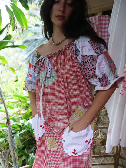A woman wearing a bohemian-style patchwork dress with red and white gingham, floral prints, and white ruffled trim, standing in a lush green outdoor setting.