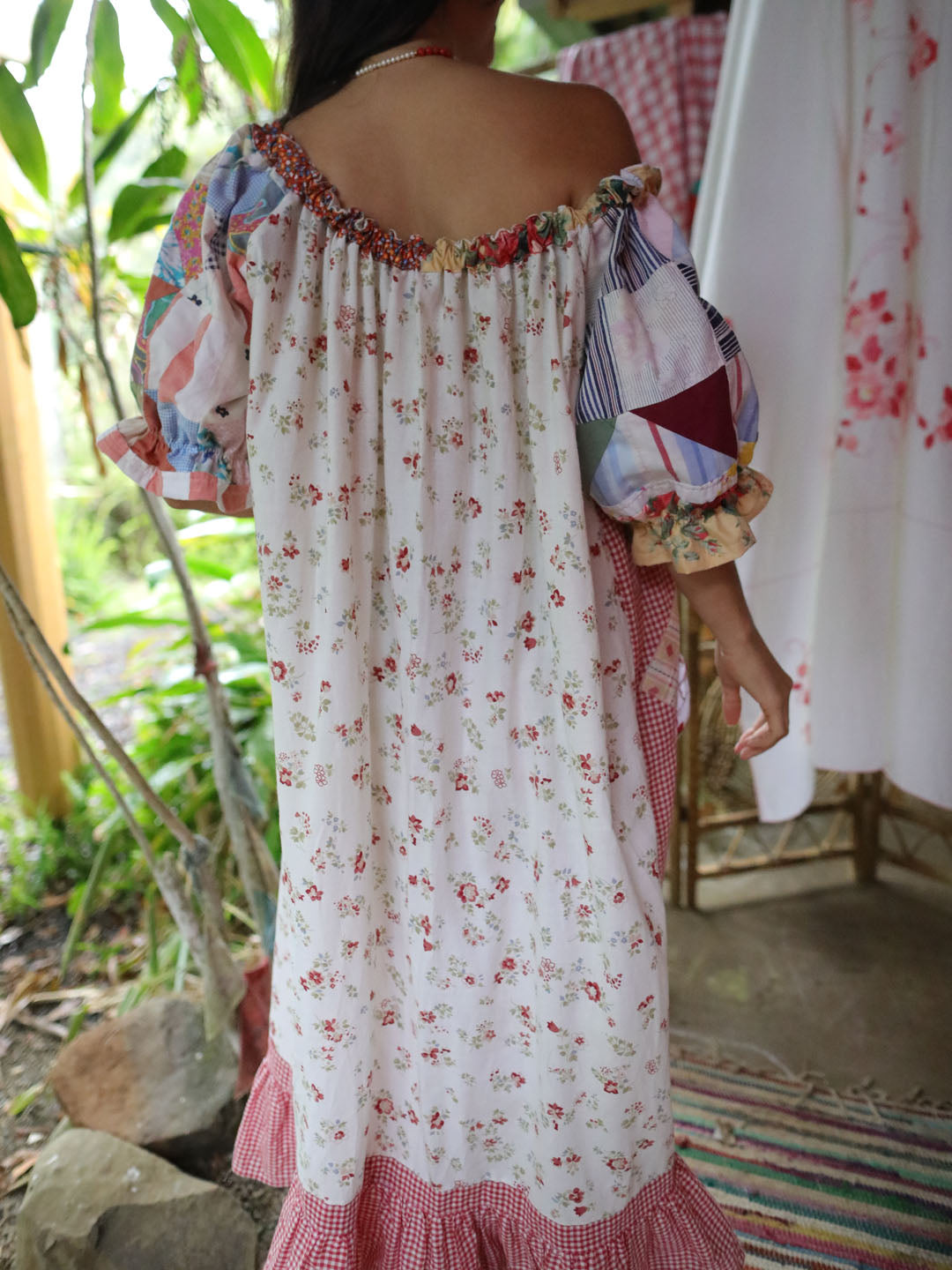 A woman wearing a bohemian-style off-the-shoulder dress with a white base, red floral print, and multicolored patchwork puff sleeves, photographed in an outdoor garden setting.