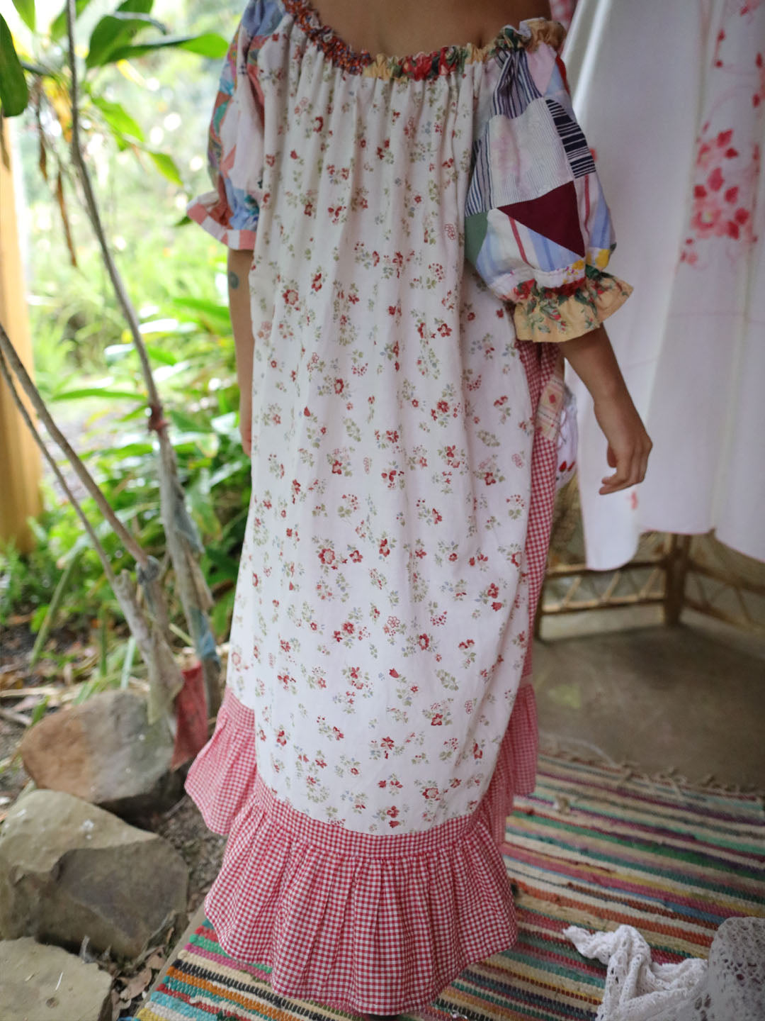 A woman wearing a bohemian-style dress with a white base, red and green floral print, patchwork puff sleeves, and a pink gingham ruffle hem, standing in a rustic garden setting.