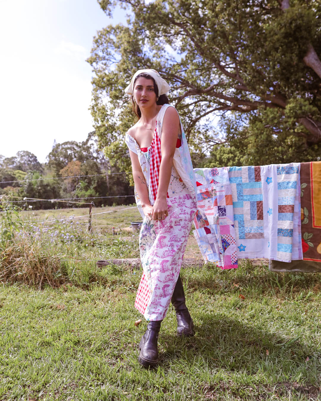 A woman stands outdoors on a grassy lawn wearing a white dress with red and pink floral patterns, a red and white gingham top, and dark boots. A colorful patchwork quilt is hanging on a clothesline in the background among trees.