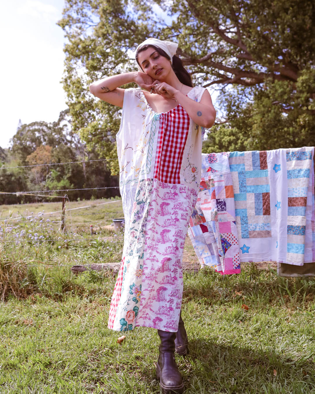 A woman stands outdoors in a grassy yard wearing a bohemian-style outfit consisting of a red and white gingham top, a white vest, a pink and white patterned skirt, and a white headscarf. A colorful patchwork quilt is hanging on a clothesline in the background with trees and a fence visible.
