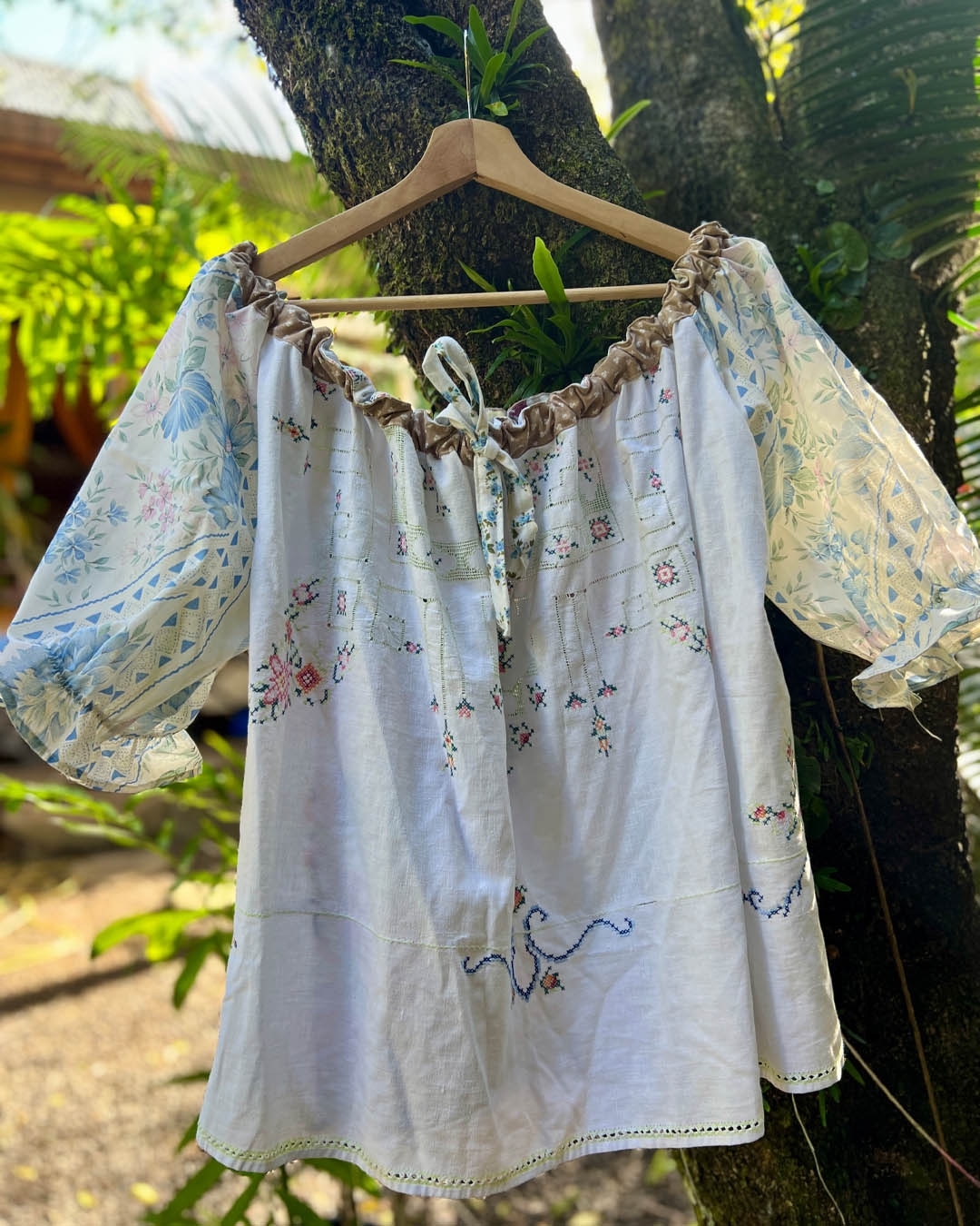 A white off-the-shoulder one size plus size smock blouse with delicate blue and pink floral embroidery heirloom fabric hanging on a wooden hanger against a tree trunk in an outdoor garden setting. 