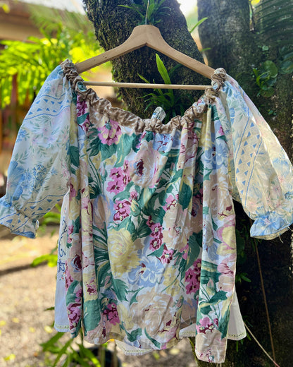 A floral off-the-shoulder one size plus size smock blouse with ruffled trim and patterned sleeves made from upcycled patchwork fabrics , hanging on a wooden hanger against a tree trunk outdoors.
