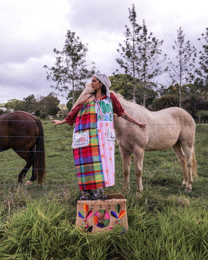 A person stands in a grassy field between two horses, wearing a long colorful plaid and patterned dress with a white headscarf and dark shoes. A woven basket sits on the grass in front of them.