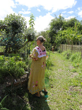 Load image into Gallery viewer, Woman in a yellow dress standing in a garden with trees and a wooden fence in the background.