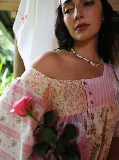 A woman wearing a pink and floral patchwork off-the-shoulder top, holding a pink rose. She is wearing a beaded necklace and hoop earrings, with a white curtain and green foliage in the background.