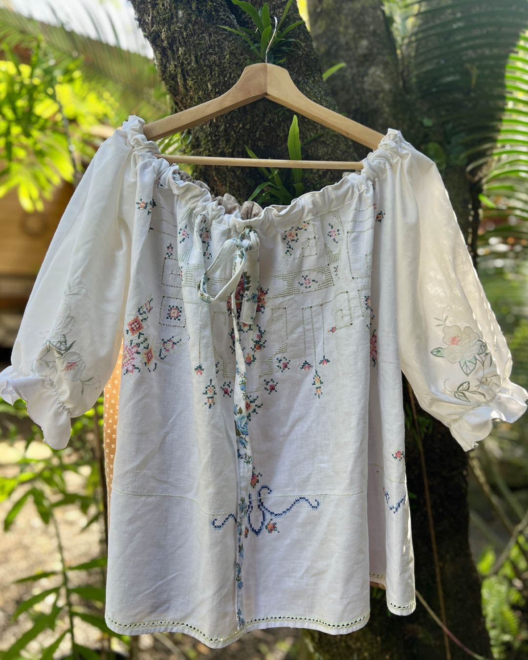 White embroidered one size plus size smock blouse with puff sleeves and a drawstring neckline, hanging on a wooden hanger against a tree trunk outdoors. Patchwork heirloom and recycled fabrics 