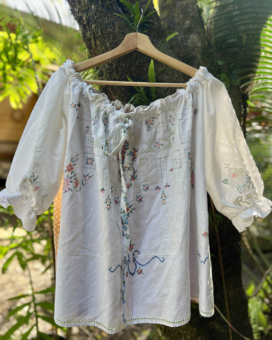 White embroidered one size plus size smock blouse with puff sleeves and a drawstring neckline, hanging on a wooden hanger against a tree trunk outdoors. Patchwork heirloom and recycled fabrics 