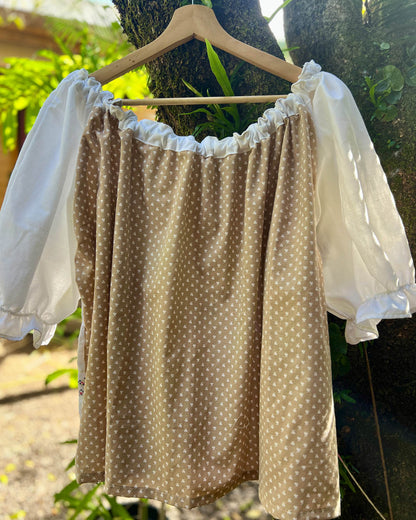 A one size plus size smock top made with vintage heirloom  brown and white polka fabric with white puff embroidered sleeves hanging on a wooden hanger outdoors against a tree trunk.