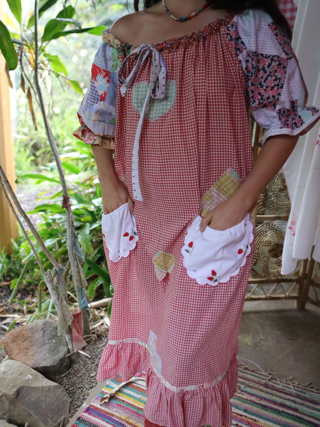 A woman wearing a red and white gingham patchwork dress with floral puff sleeves, white scalloped pockets with strawberry embroidery, and a ruffled hem, standing in a garden setting.