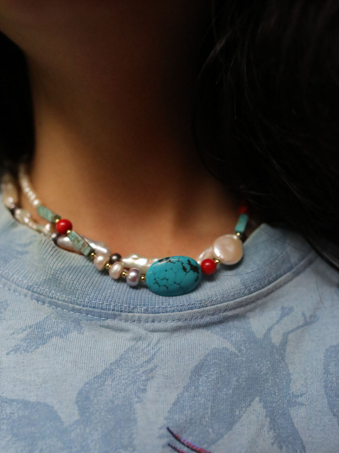 A close-up of a person wearing a beaded necklace with a prominent turquoise stone, white pearls, and red beads, paired with a light blue patterned t-shirt.