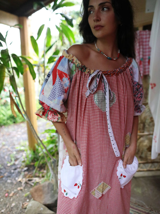 A woman wearing a red and white gingham patchwork dress with floral puff sleeves, a tie neckline, and white pockets, standing outdoors on a porch with greenery in the background.