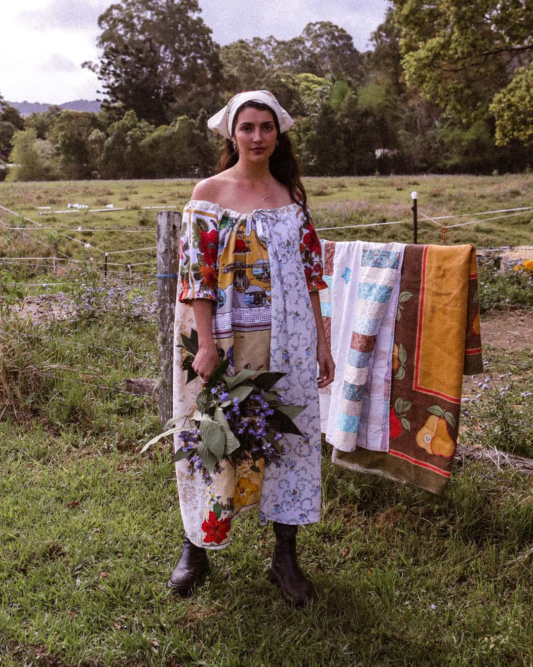 A woman stands outdoors in a grassy field wearing a colorful off-the-shoulder patchwork dress and holding a bouquet of purple flowers. A matching patchwork quilt is hanging on a fence behind her.