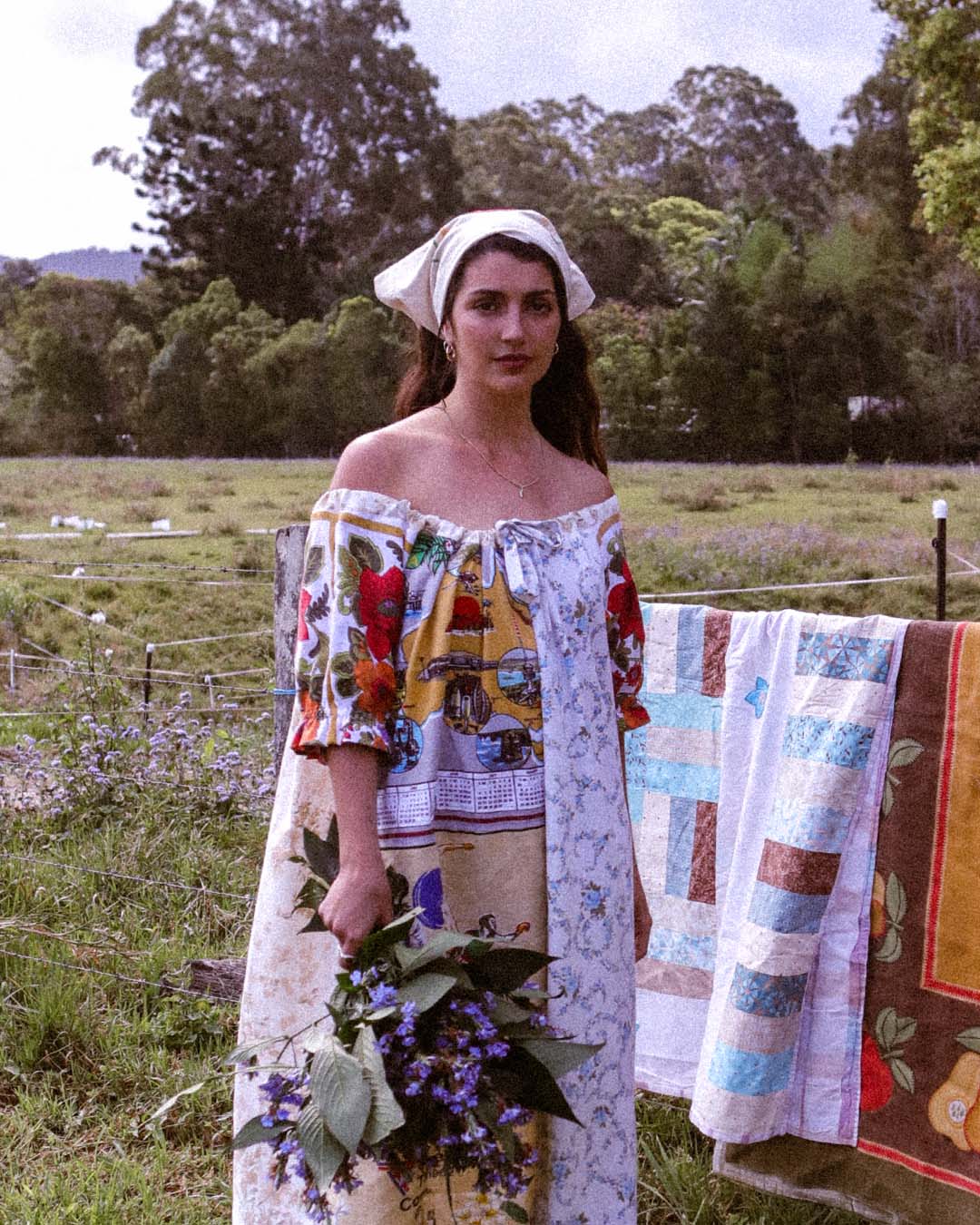 A woman stands outdoors in a grassy field wearing a colorful off-the-shoulder dress with a white headscarf, holding a bouquet of purple flowers. A quilted blanket hangs on a fence behind her, with trees in the background.