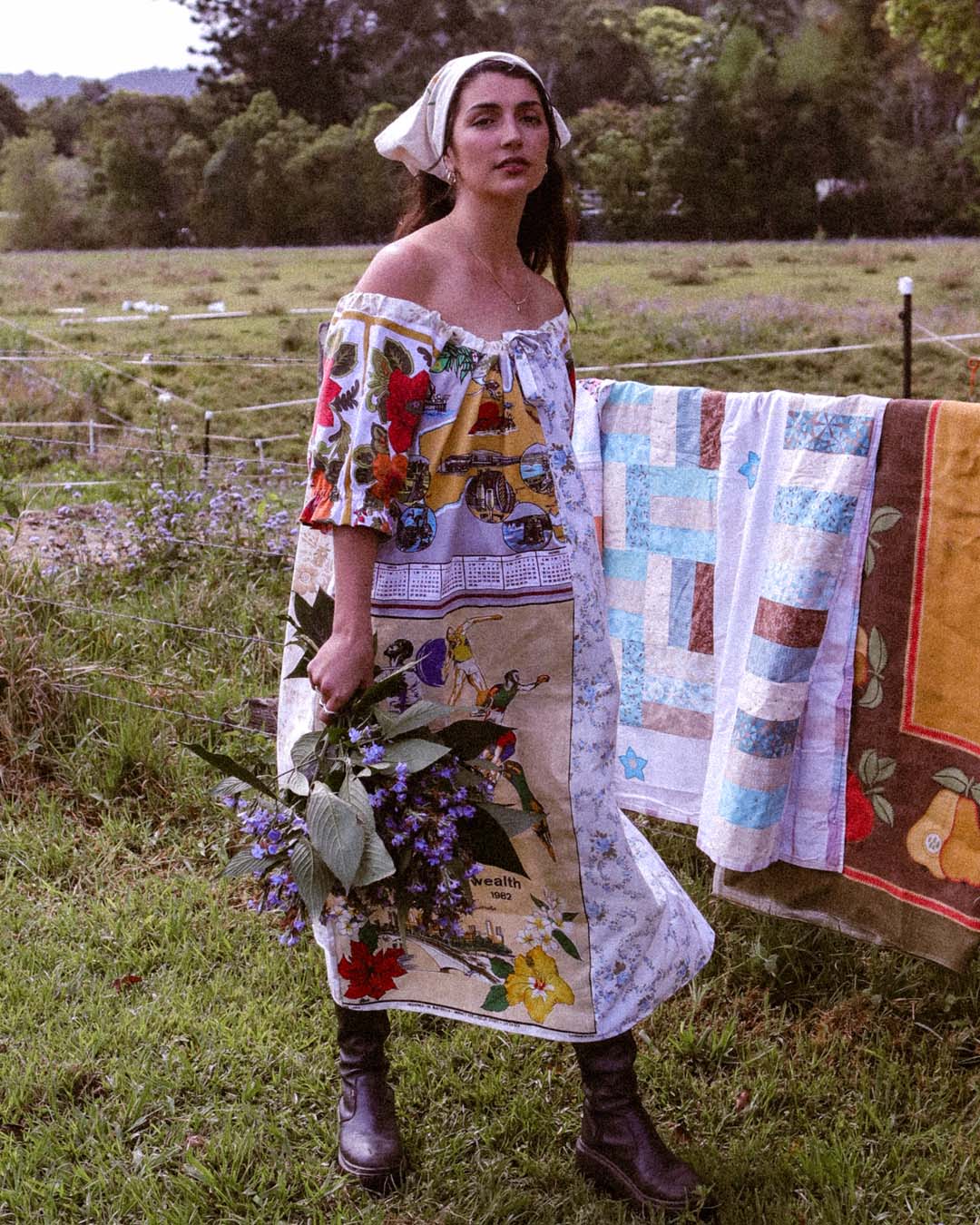 A woman stands outdoors in a grassy field wearing a colorful off-the-shoulder patchwork dress with floral and vintage-style prints, holding a bouquet of purple flowers. A quilted blanket is draped over a fence behind her.