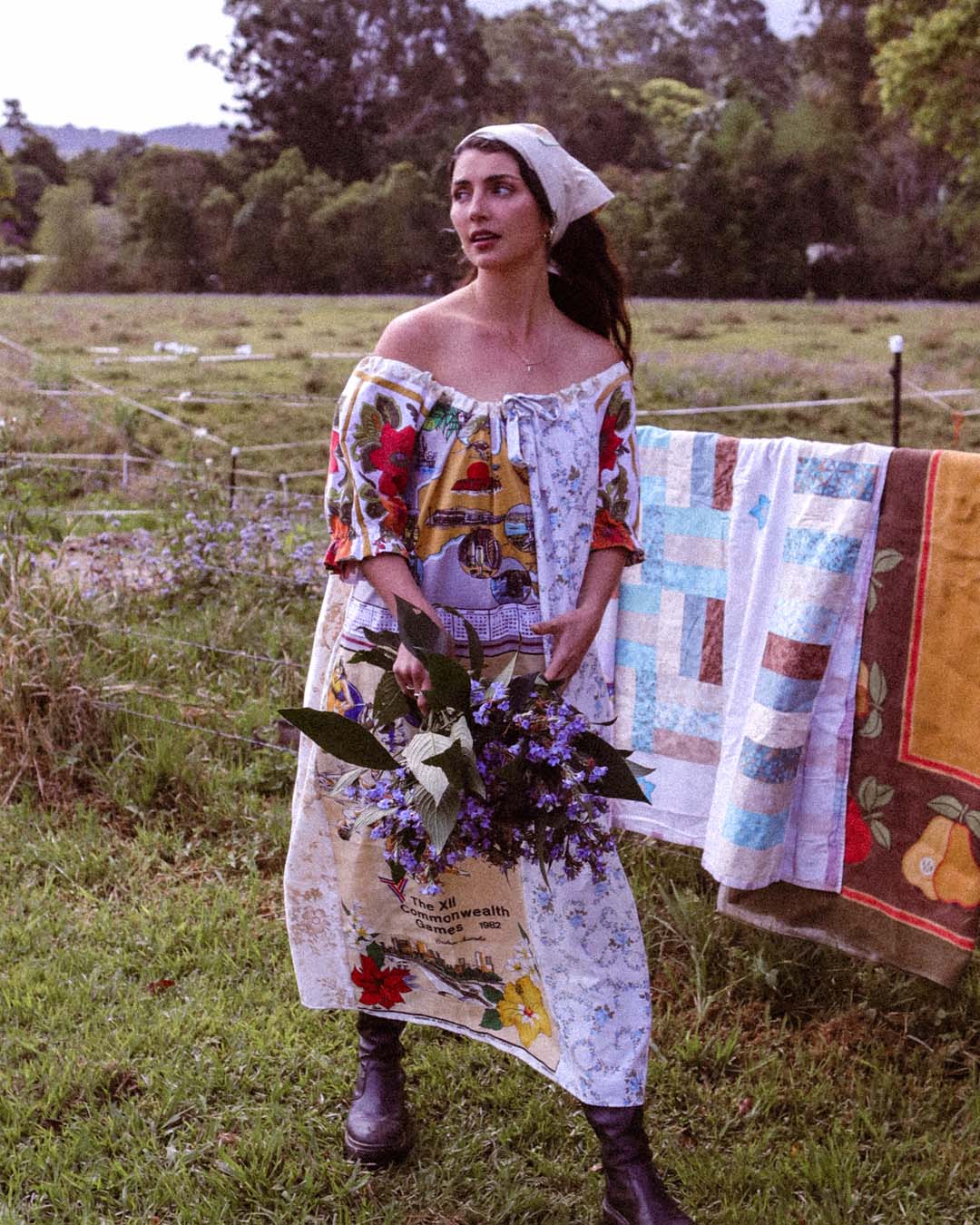 A woman stands in a grassy field wearing a colorful off-the-shoulder patchwork dress with a white headscarf and dark boots, holding a bouquet of purple flowers. A quilted blanket hangs on a fence behind her.