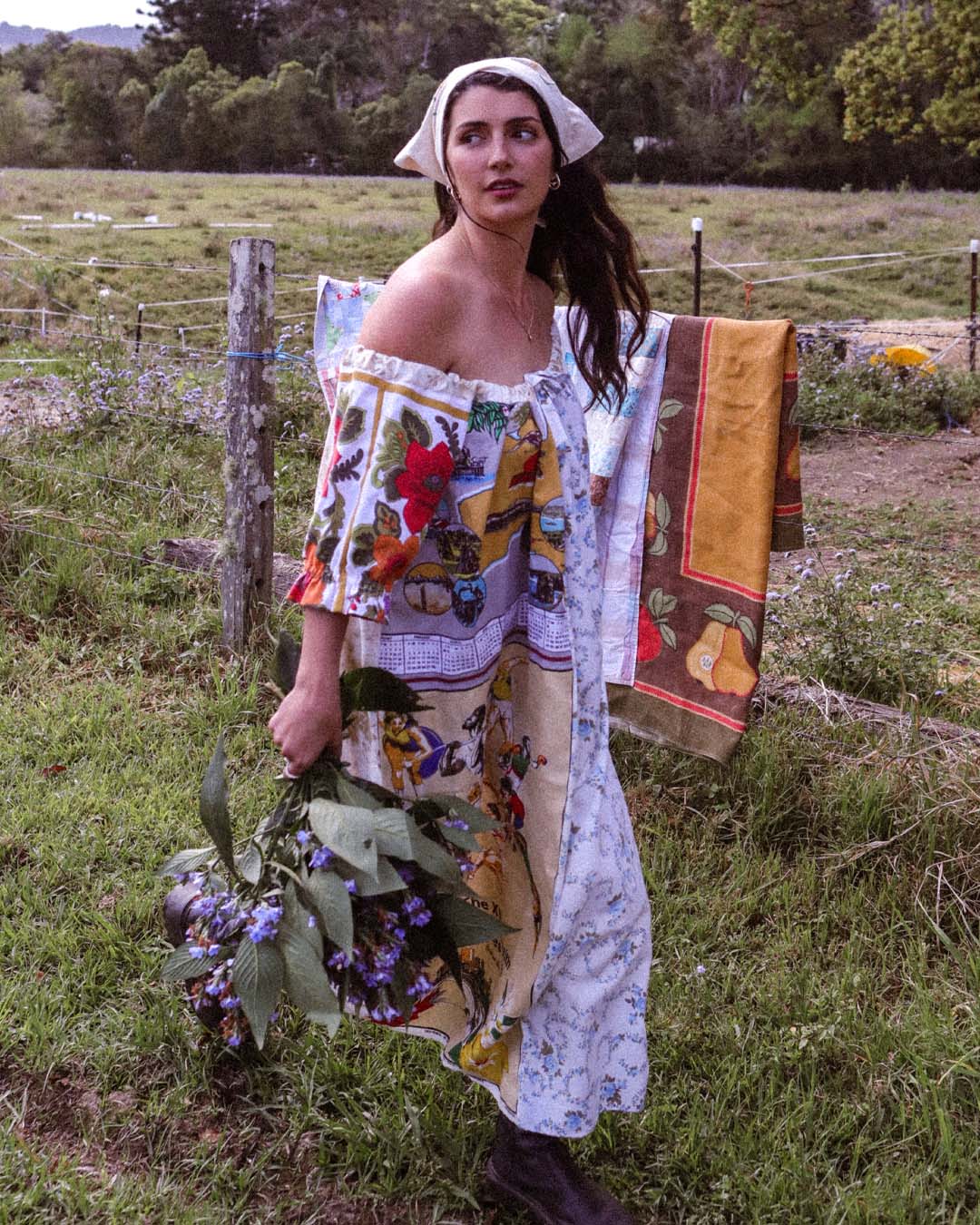 A woman stands outdoors in a grassy field wearing a colorful off-the-shoulder dress made from patchwork fabrics, with a white headscarf and dark boots. She holds a bouquet of green leaves and purple flowers, and a patterned textile hangs from a fence post behind her.