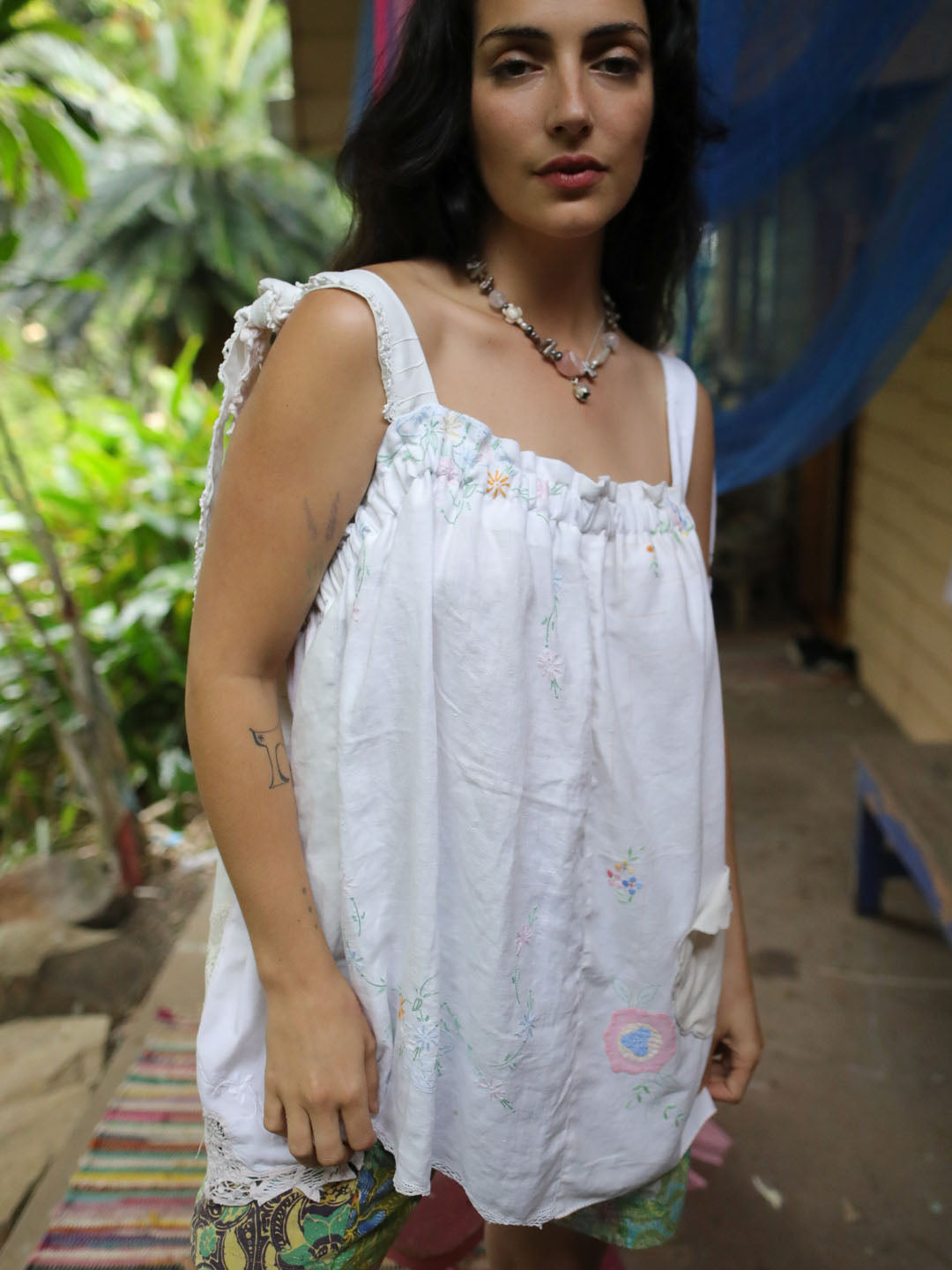 A woman wearing a white sleeveless top with delicate pastel floral embroidery and ruffled shoulder details, standing in an outdoor setting with green foliage and a rustic background.
