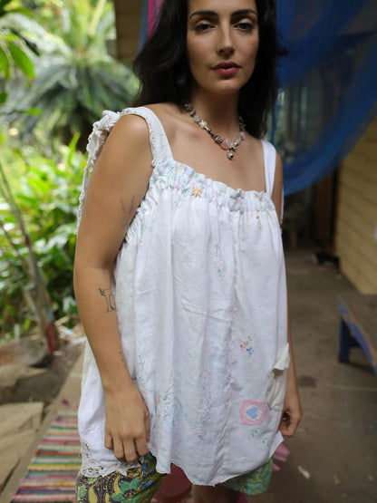 A woman wearing a white sleeveless top with delicate pastel floral embroidery and ruffled shoulder details, standing in an outdoor setting with green foliage and a rustic background.
