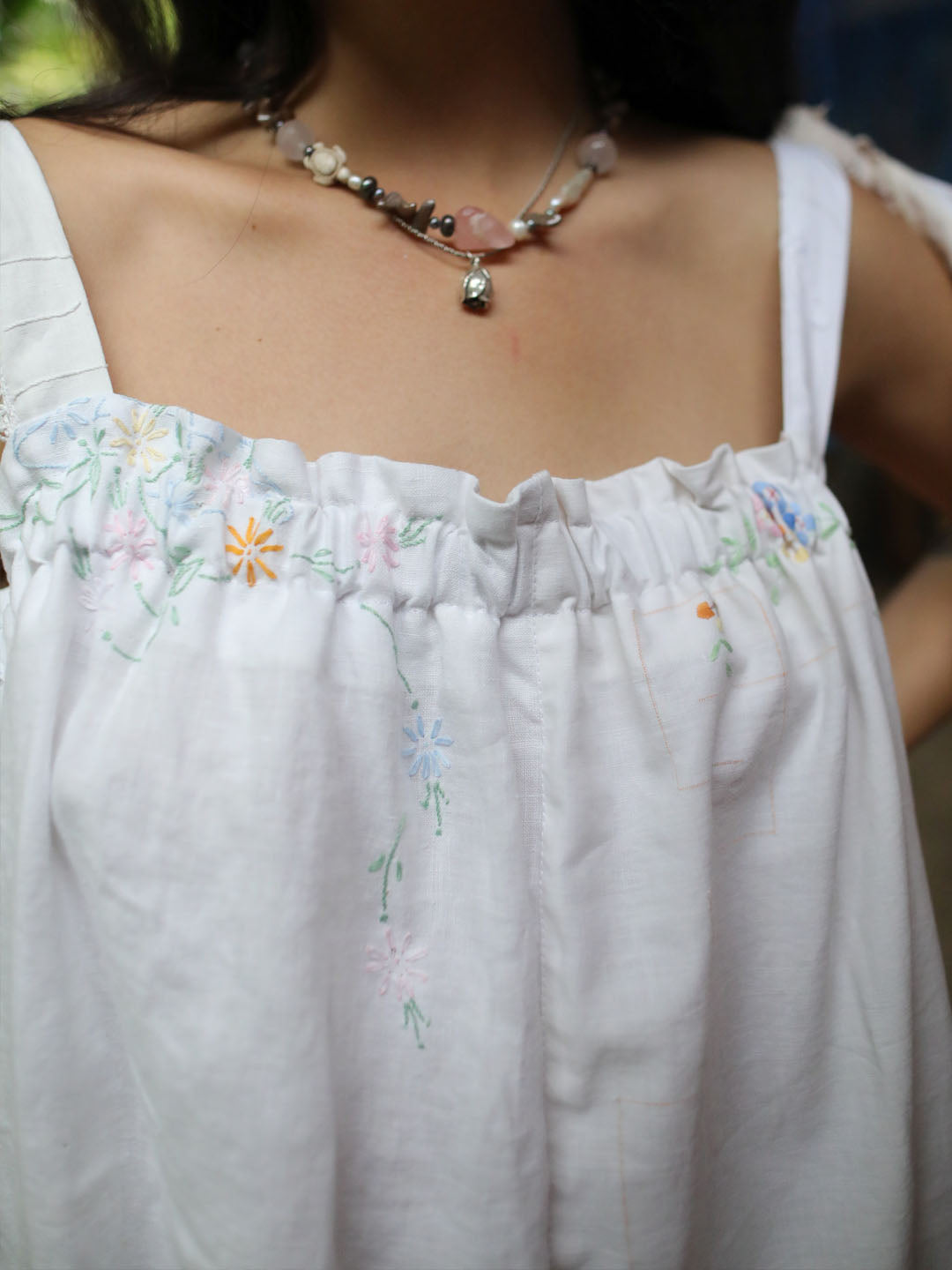 A close-up of a person wearing a white embroidered top with delicate pastel floral stitching and a beaded necklace.