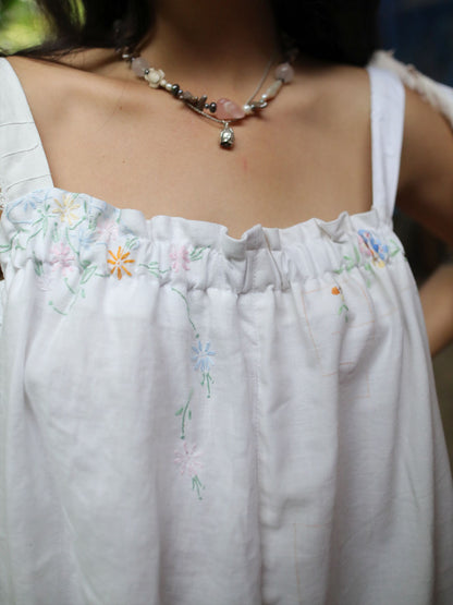 A close-up of a person wearing a white embroidered top with delicate pastel floral stitching and a beaded necklace.