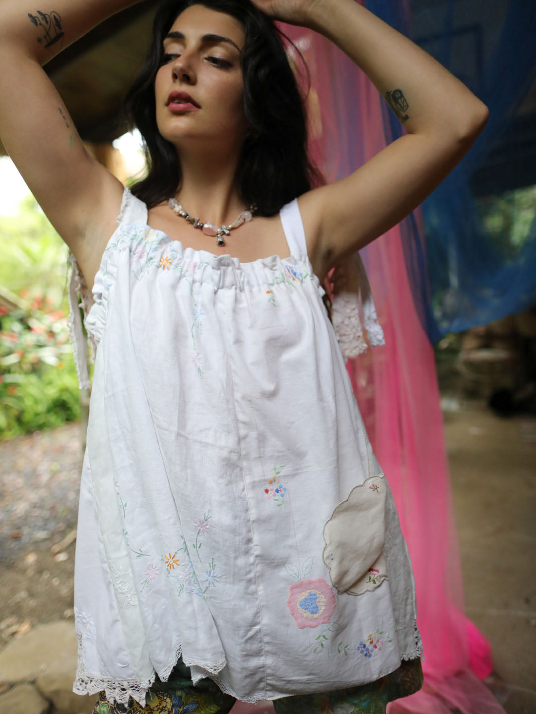 A woman wearing a white sleeveless embroidered top with lace trim and a patch detail, posed outdoors with colorful fabric drapes in the background.