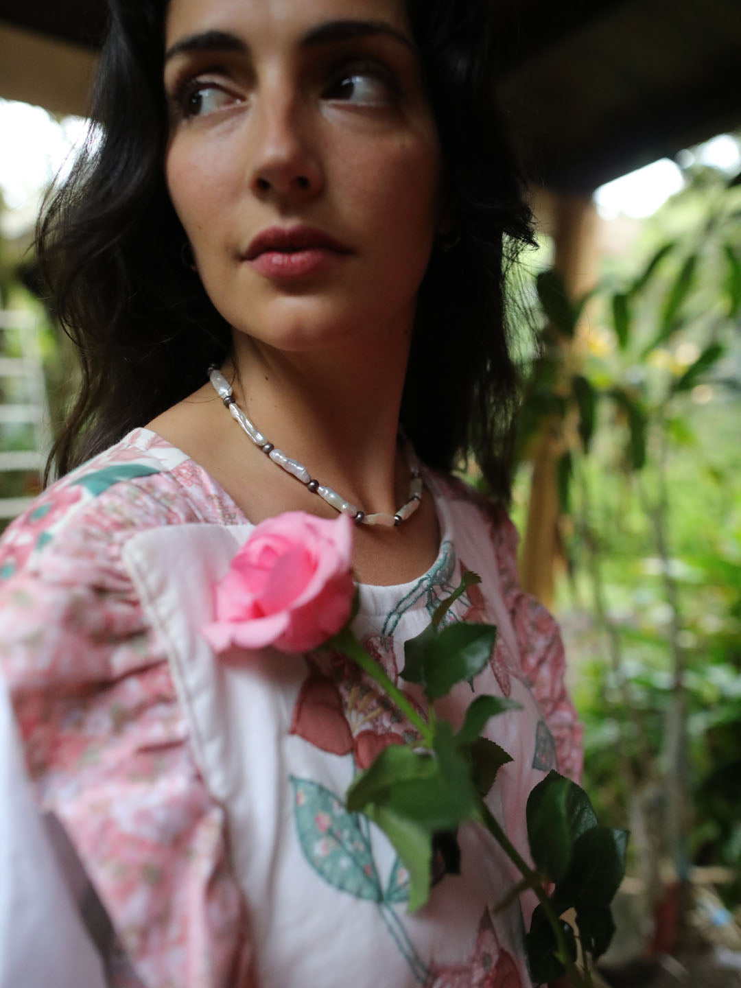 A woman wearing a light pink floral print top and a silver beaded necklace, holding a pink rose in a garden setting.