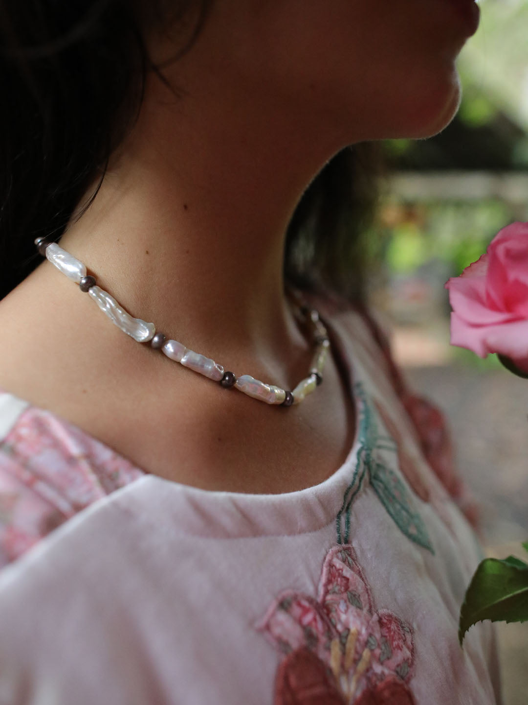 A close-up lifestyle shot of a woman wearing a handmade beaded necklace made of white baroque pearls and small dark metallic beads, paired with a light pink floral embroidered top.