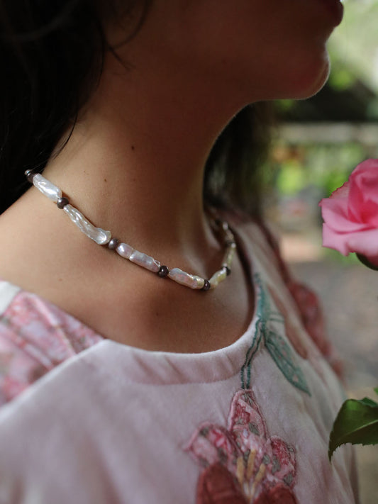 A close-up lifestyle shot of a woman wearing a handmade beaded necklace made of white baroque pearls and small dark metallic beads, paired with a light pink floral embroidered top.