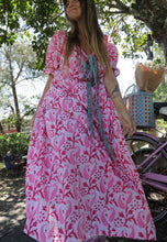 Load image into Gallery viewer, Woman in a pink floral dress standing outdoors with a bicycle and basket in the background.