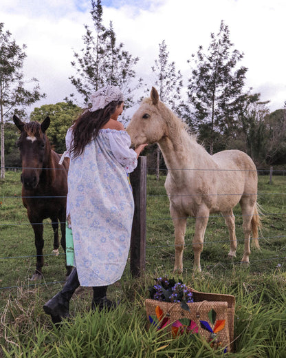 A woman in a light blue floral dress and bonnet stands in a grassy field, touching the face of a light-colored horse while another dark horse stands behind her. A woven basket with colorful flowers sits on the ground in the foreground.