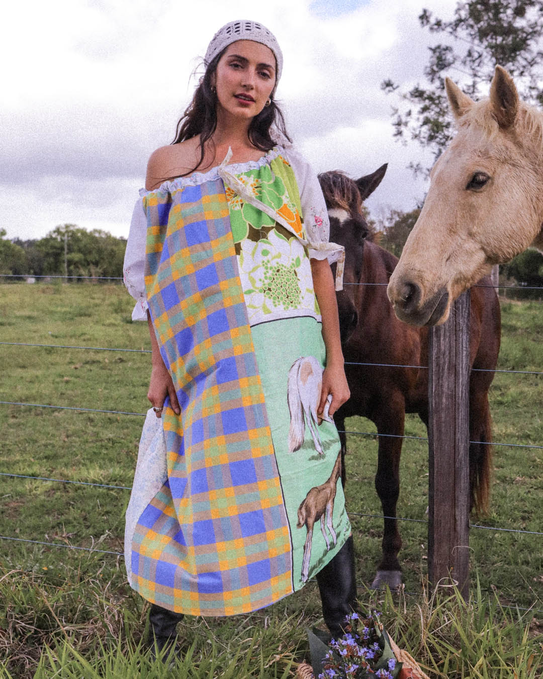 A woman wearing a colorful one size one of a kind patchwork dress stands in a grassy field next to a brown horse and a white horse, with a white sky in the background.