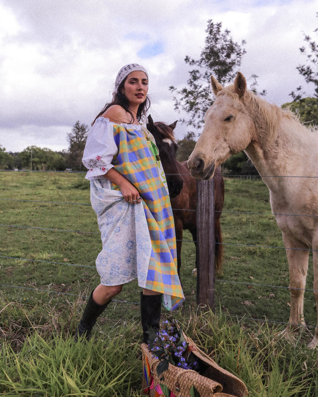 A woman wearing a white off-the-shoulder dress with floral embroidery and a blue, yellow, and purple checkered apron stands in a grassy field next to a white horse and a brown horse, with a basket in the foreground.