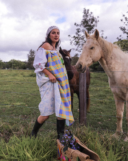 A woman wearing a white off-the-shoulder dress with floral embroidery and a blue, yellow, and purple checkered apron stands in a grassy field next to a white horse and a brown horse, with a basket in the foreground.