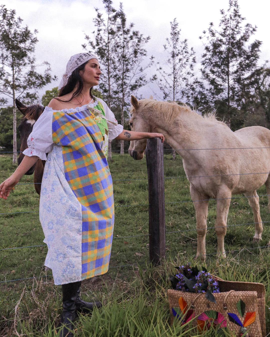 A woman wearing a colorful patchwork dress stands in a grassy field next to a light-colored horse, with trees and an overcast sky in the background.
