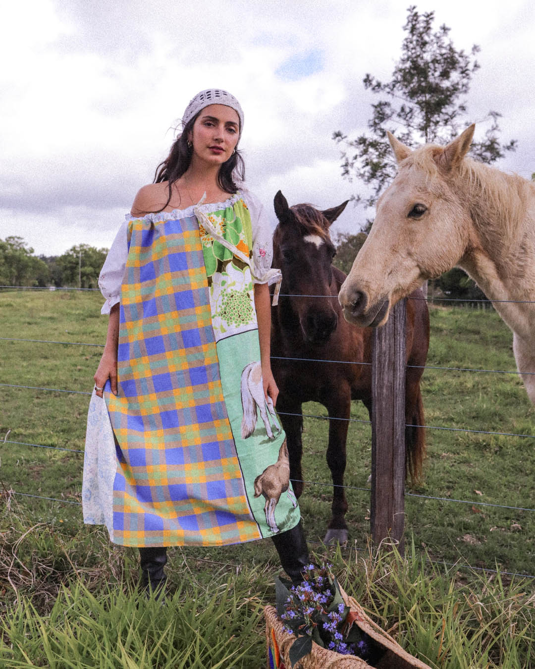 A woman stands in a grassy field with two horses behind a wire fence, wearing a colorful patchwork apron dress with blue, yellow, green, and white plaid sections and animal illustrations. A basket of flowers sits on the ground in the foreground.